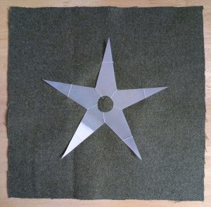 A plastic, star shaped template with a circle cut out of the middle. It is basted to a square of wool fabric using sewing thread and large stitches.