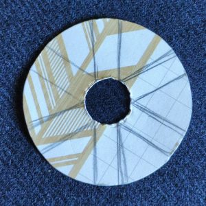 A two inch circle of cardboard, with a half inch hole cut out of the middle. It has six thick lines radiating out from the centre at regular intervals. It sits flat on a piece of woollen fabric.