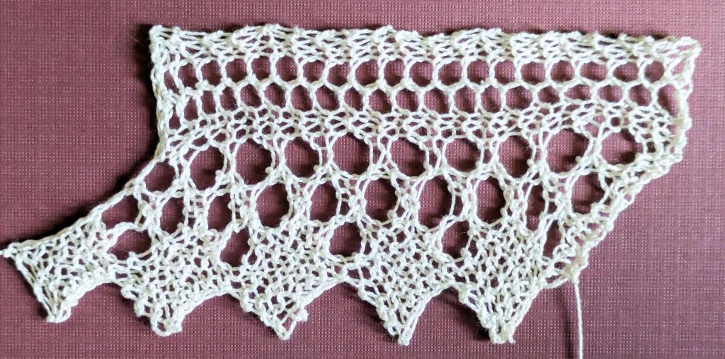 Wide knitted lace with a sawtooth edge made of solid diamonds. Above the diamonds is a triangular design made of large eyelets.
