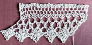 Wide knitted lace with a sawtooth edge made of solid diamonds. Above the diamonds is a triangular design made of large eyelets.